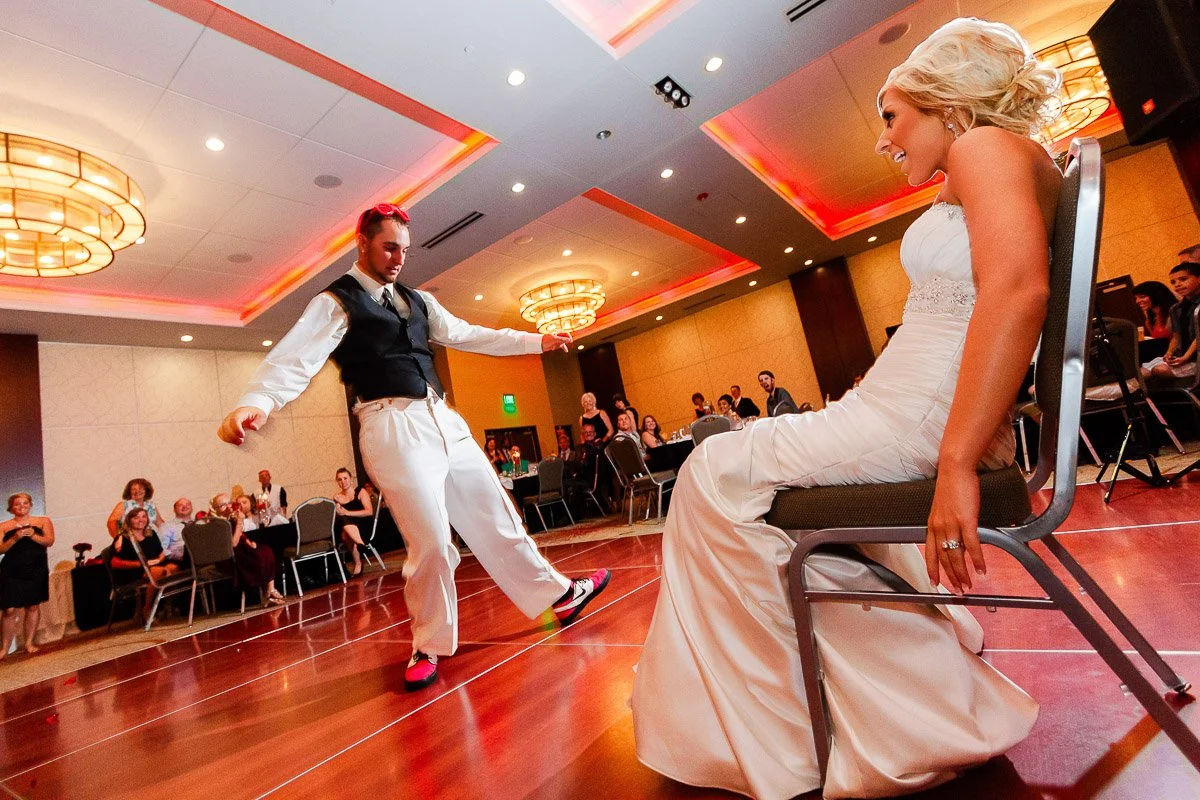 A bride in a white gown smiles and sits on a chair, watching a colorful shoe-wearing groom dance energetically on a ballroom floor. Guests watch and laugh.