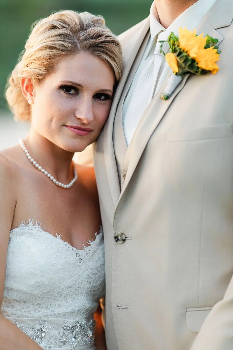 A joyous couple stands outdoors beside a serene pond during sunset, with lush greenery in the background. They are dressed in elegant wedding attire.