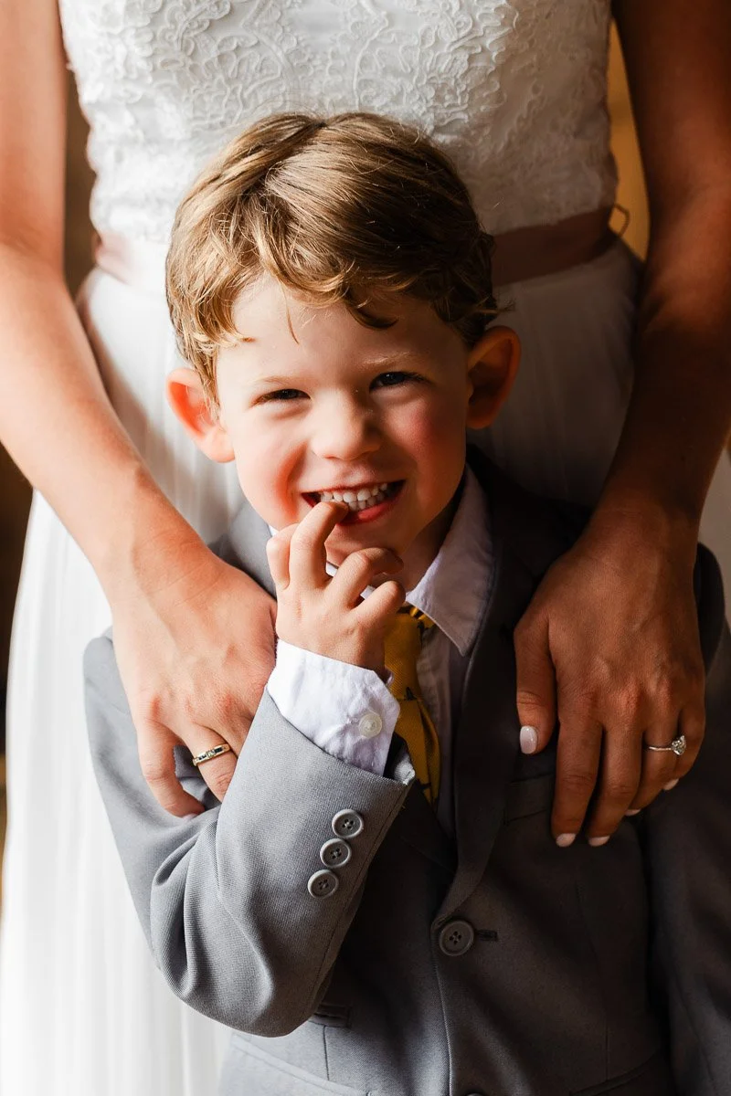 A smiling young boy in a gray suit with a yellow tie stands in front of an adult, whose arms gently wrap around him, expressing warmth and affection.