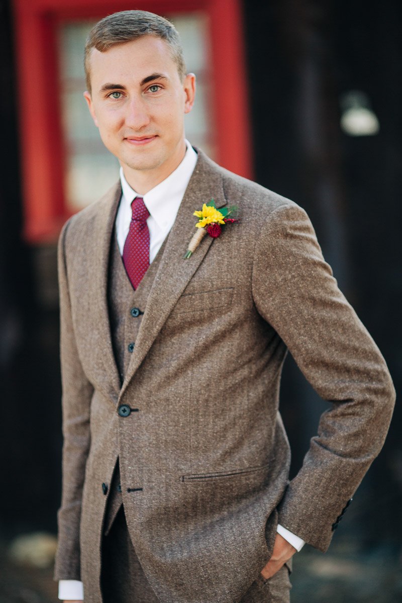 A man in a brown tweed suit with a red tie and a flower boutonniere smiles subtly. The background is blurred, suggesting either a natural or rustic setting.
