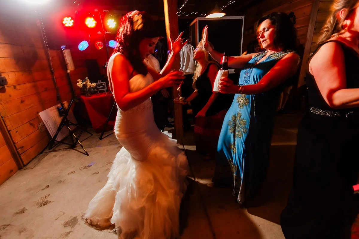 Bride in a white gown and guests dance energetically at a wedding reception. Warm lights and wooden walls create a festive, joyful atmosphere.