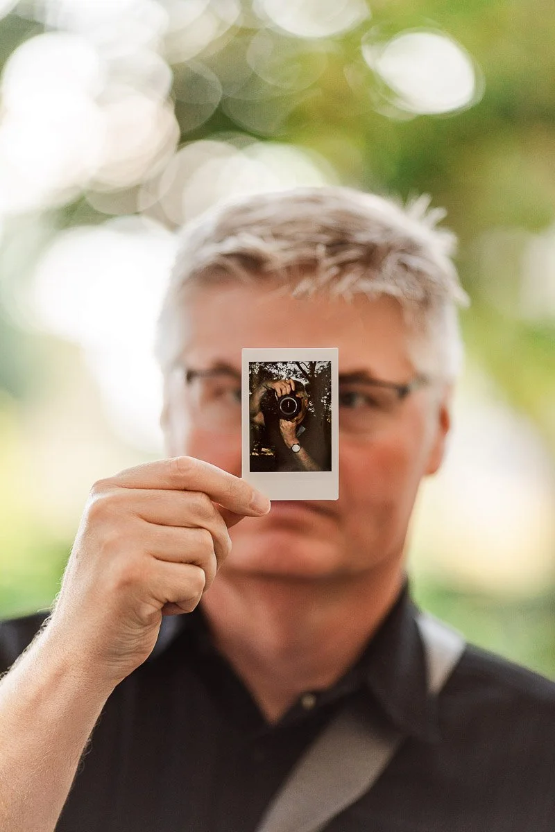 A person with light hair holds a small photo in front of their face, creating a playful effect. The background is softly blurred green foliage.