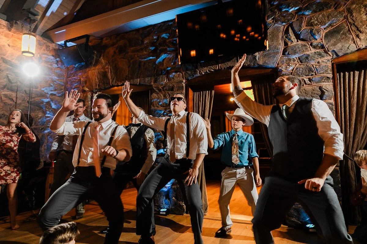 A group of people dressed in formal attire energetically dance indoors. The stone walls and dim lighting create a festive atmosphere.