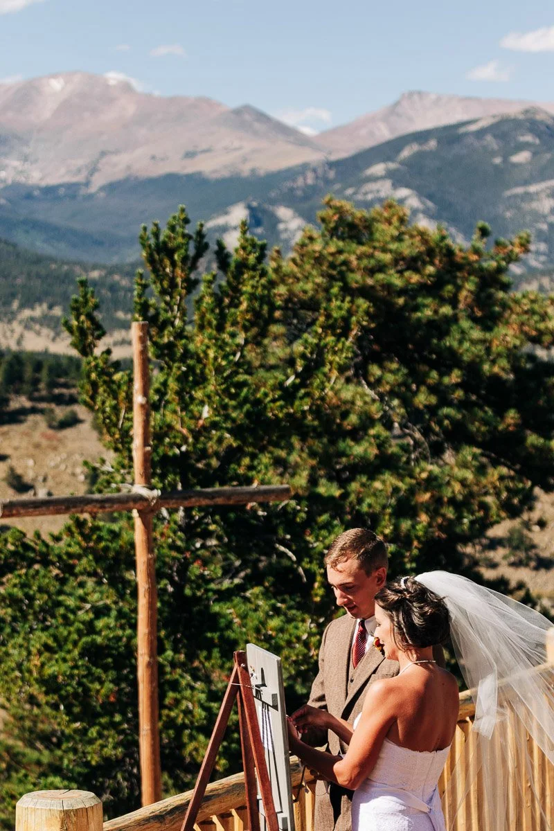 A bride and groom stand by an easel outdoors during a wedding ceremony. Behind them, a wooden cross and mountainous landscape create a serene backdrop.