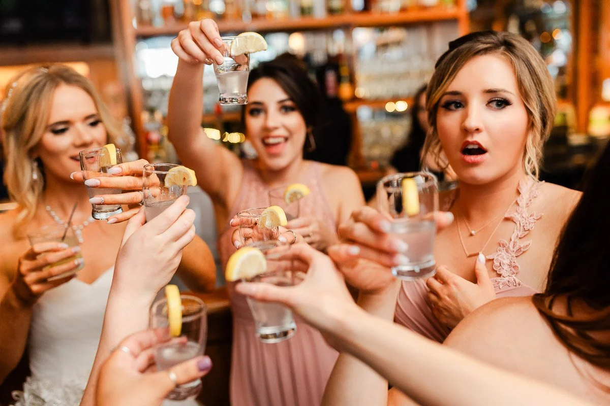 Women in pastel dresses joyfully toast with lemon-garnished drinks, conveying celebration and camaraderie in a lively bar setting during a The Lobby wedding reception in Denver.