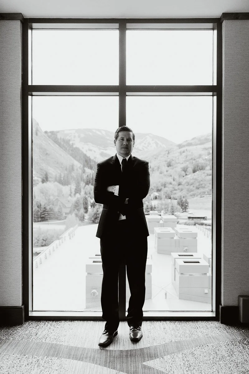 Man in a suit stands confidently in front of a large window with a mountainous landscape visible outside. The mood is professional and serene.