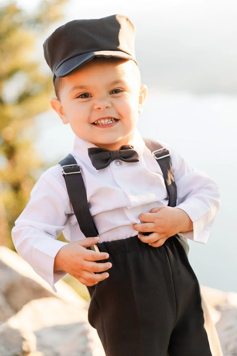 A smiling young boy wearing a classic black cap, bow tie, and suspenders on a white shirt. He stands outdoors, with sunlight creating a joyful atmosphere.