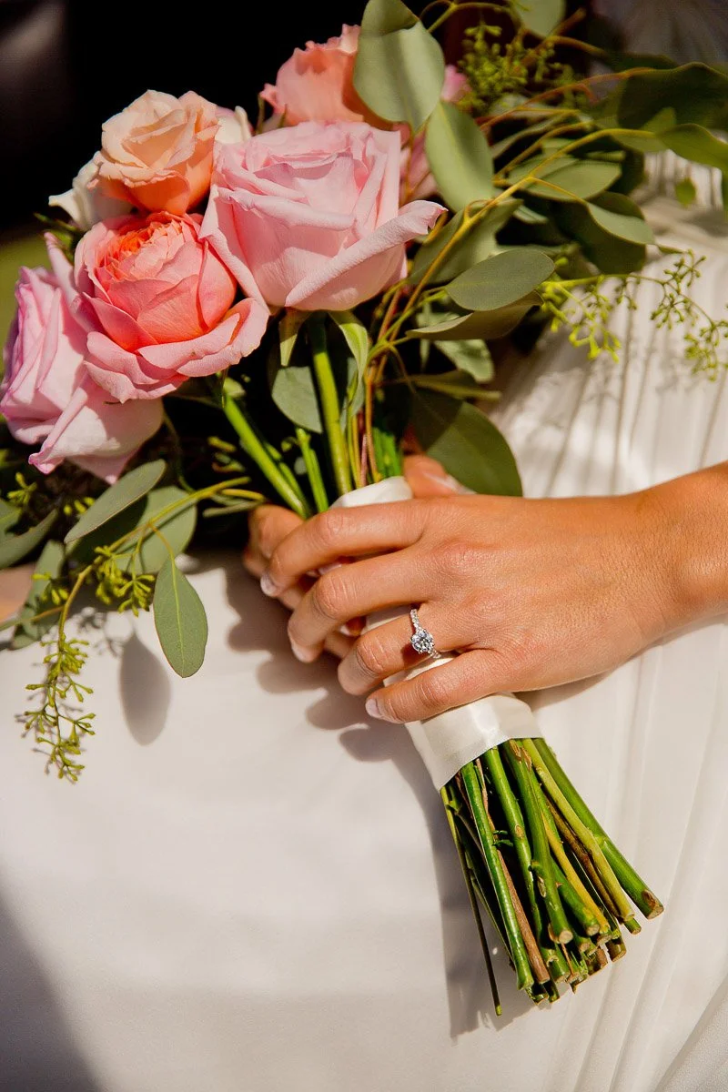 A hand holding a bouquet of pink roses and greenery, wrapped in white ribbon. A diamond ring is visible, conveying a joyful, romantic tone.