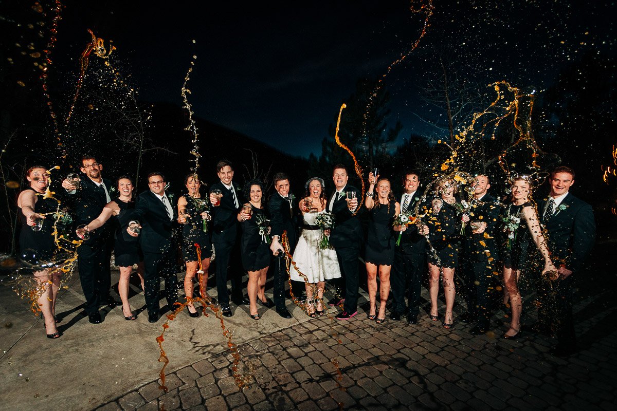 A joyful wedding party poses outdoors at night, laughing as they spray champagne. The group is dressed in formal attire, and the scene is festive and lively.