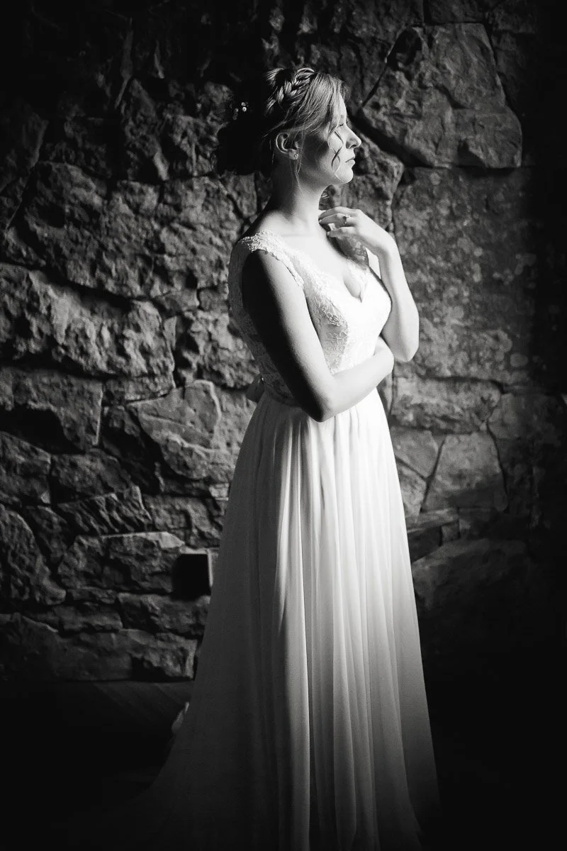 A woman in a white lace gown stands in profile, gazing thoughtfully. Stone wall in the background. Soft lighting creates a serene, contemplative mood.