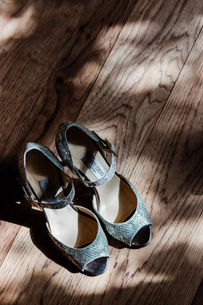 Elegant, glittery high-heeled shoes with ankle straps sit on a sunlit wooden floor, creating a contrast of light and shadow.