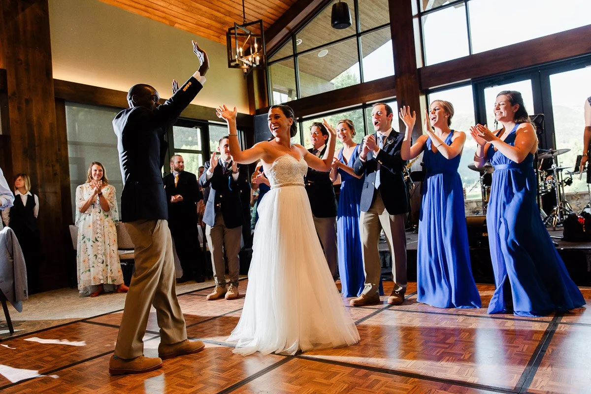 A joyful bride in a white dress dances with a groomsman on a wooden floor, surrounded by cheering bridesmaids in blue dresses and groomsmen in suits.