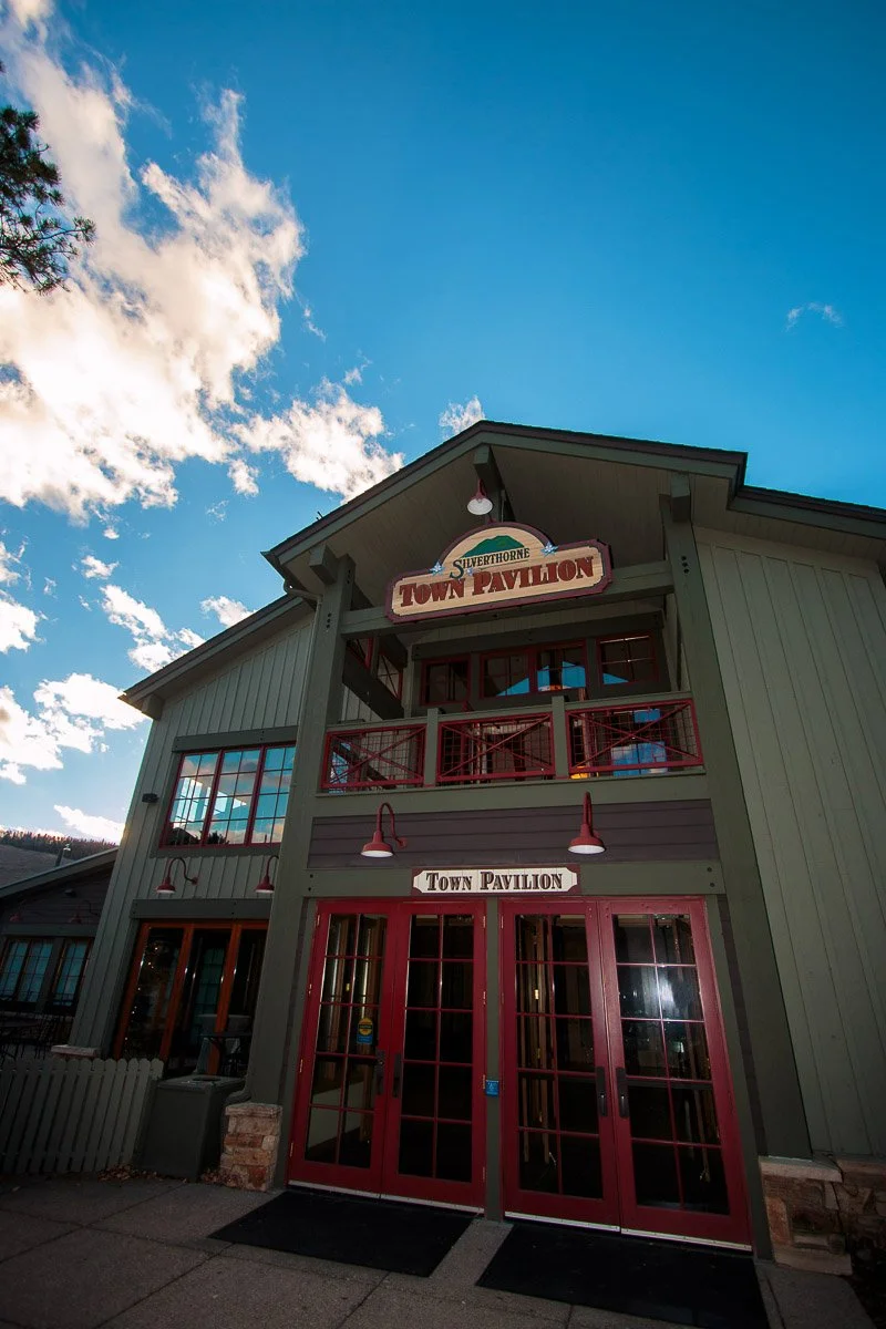 Silverthorne Pavilion building under a vibrant blue sky with scattered clouds. The structure features red doors and trim, creating a welcoming, rustic appearance.