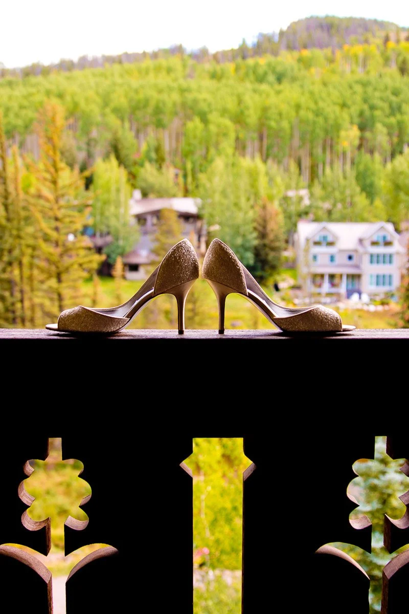 Elegant high heels rest on a wooden railing, framed by a lush green forest and distant houses, conveying a serene and sophisticated mood.