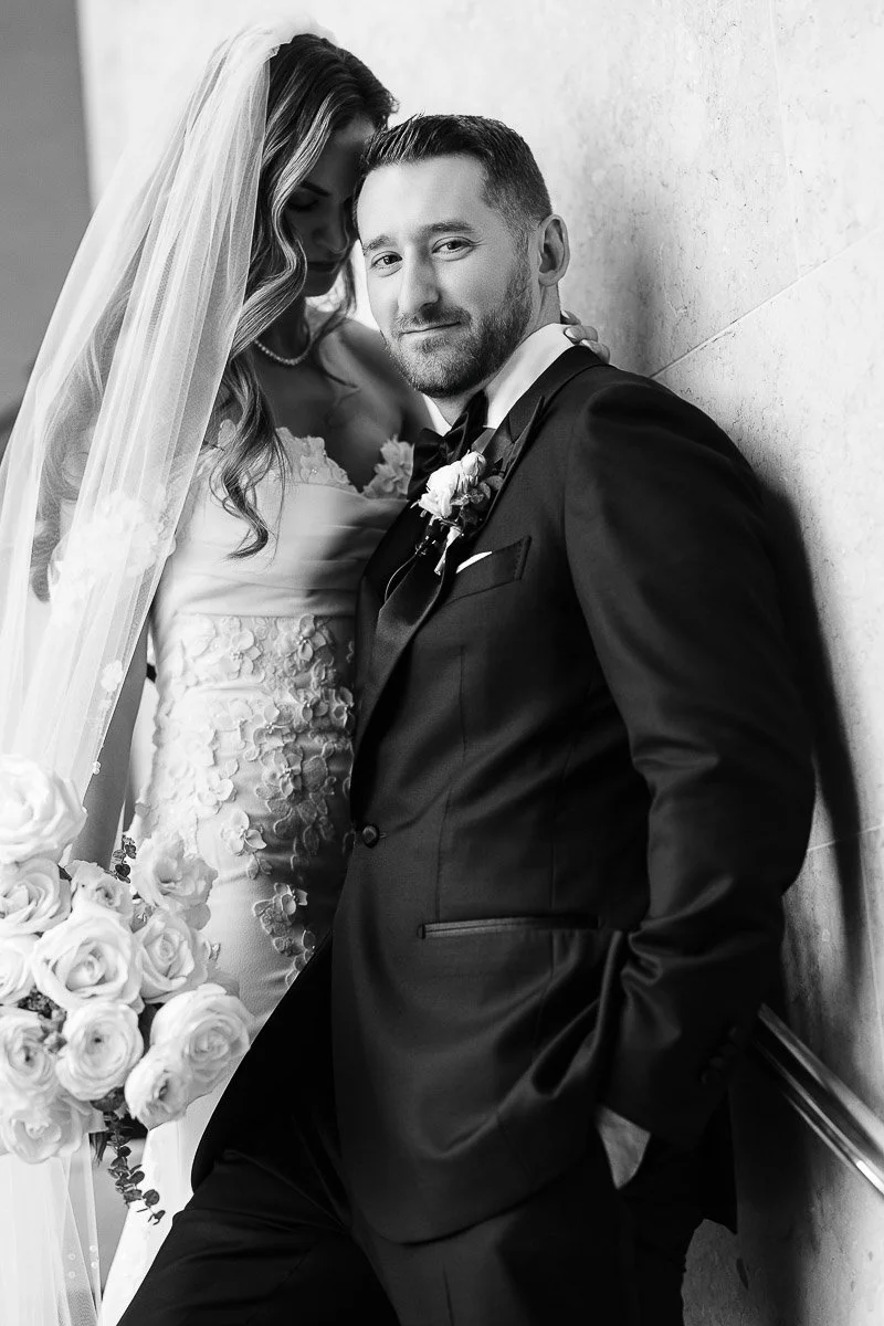 Black and white wedding photo of a smiling groom in a suit leaning against a wall, with the bride in a floral gown and veil holding roses beside him.