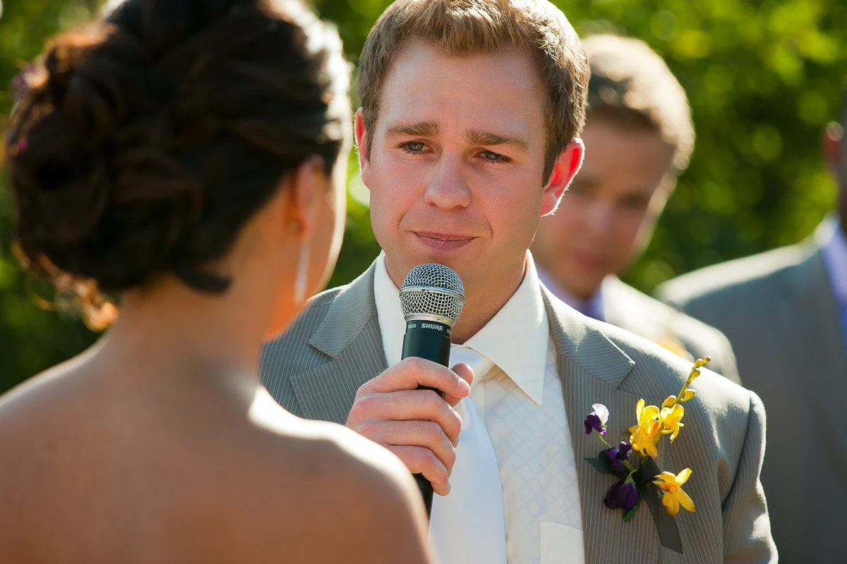 Man in a gray suit with floral boutonniere speaks emotionally into a microphone, facing a woman, during an outdoor wedding ceremony.