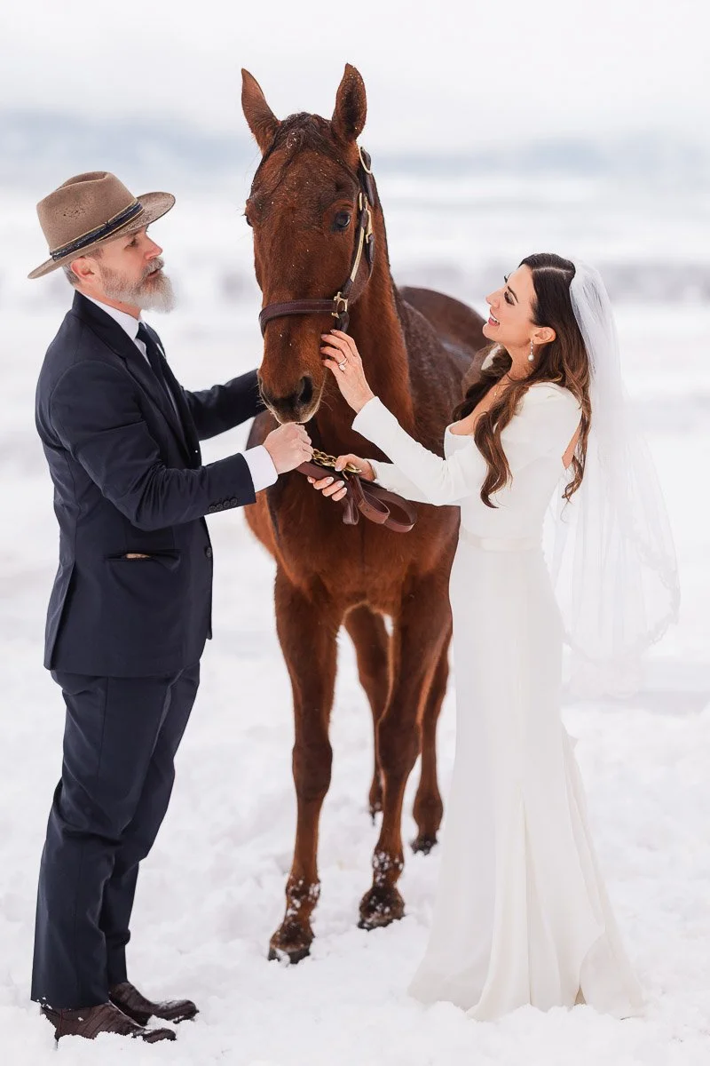 Bride in a white dress and groom in a suit stand with a brown horse in a snowy landscape. They gently hold the horse, conveying joy and serenity captured by La Joya Dulce wedding photographer tomKphoto.
