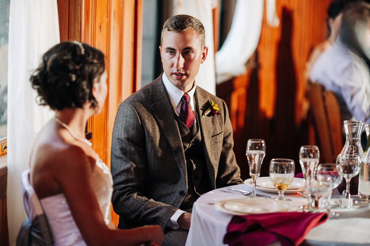 A couple, possibly at a wedding, sits at a table set with glasses and plates. The man in a suit looks at the woman, who is in a white dress, engaged in conversation.