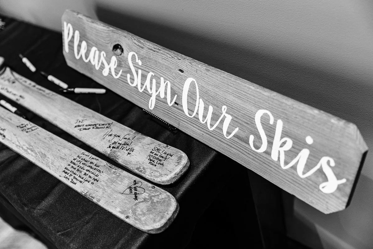 Black and white image of a wooden sign reading "Please Sign Our Skis" above two skis with handwritten messages, on a black tablecloth.