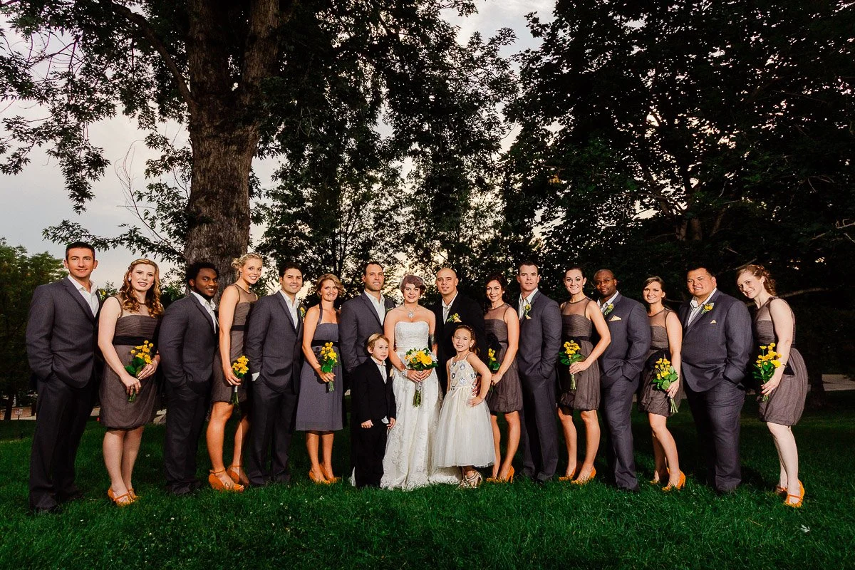 A joyful wedding party stands on grass, surrounded by trees. The bride and groom are in the center, with bridesmaids in grey dresses and groomsmen in suits.