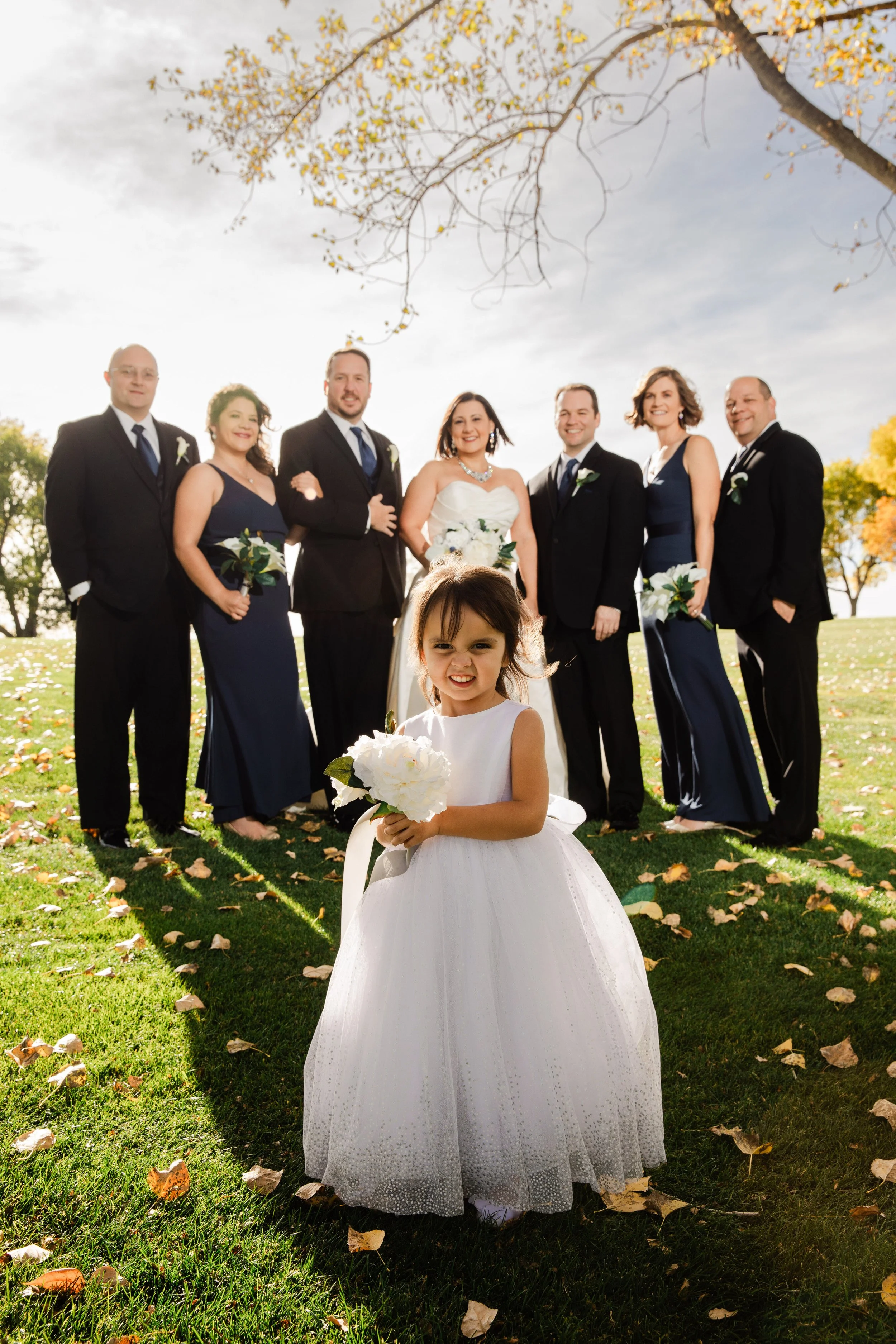 A joyful wedding scene with a smiling girl in a white dress holding flowers in the foreground. Behind her, a wedding party in formal attire stands on a grassy lawn under a partly cloudy sky.