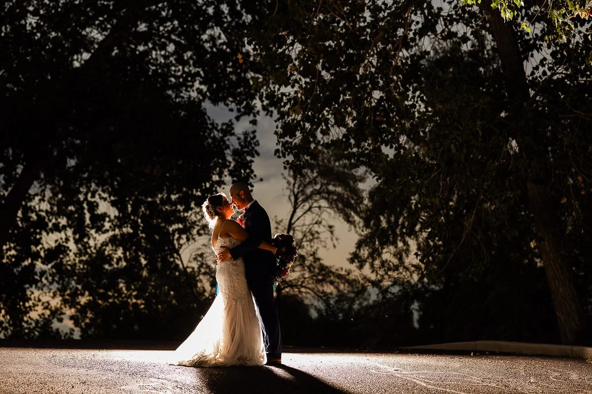 A bride and groom embrace romantically under a spotlight, surrounded by dark trees at night. The scene conveys warmth and intimacy captured by Denver wedding photographer tomKphoto