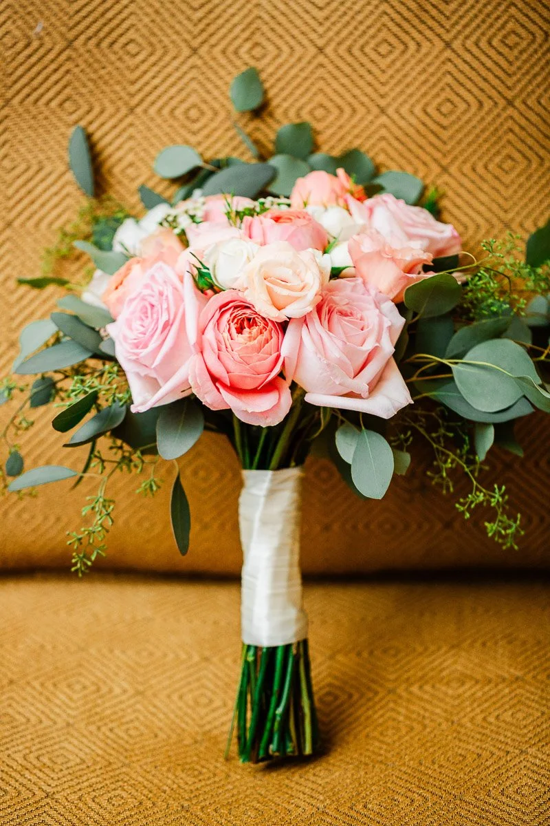 A bouquet of pink and peach roses with eucalyptus leaves, wrapped in white ribbon, set against a textured, tan geometric background. The tone is elegant and romantic.