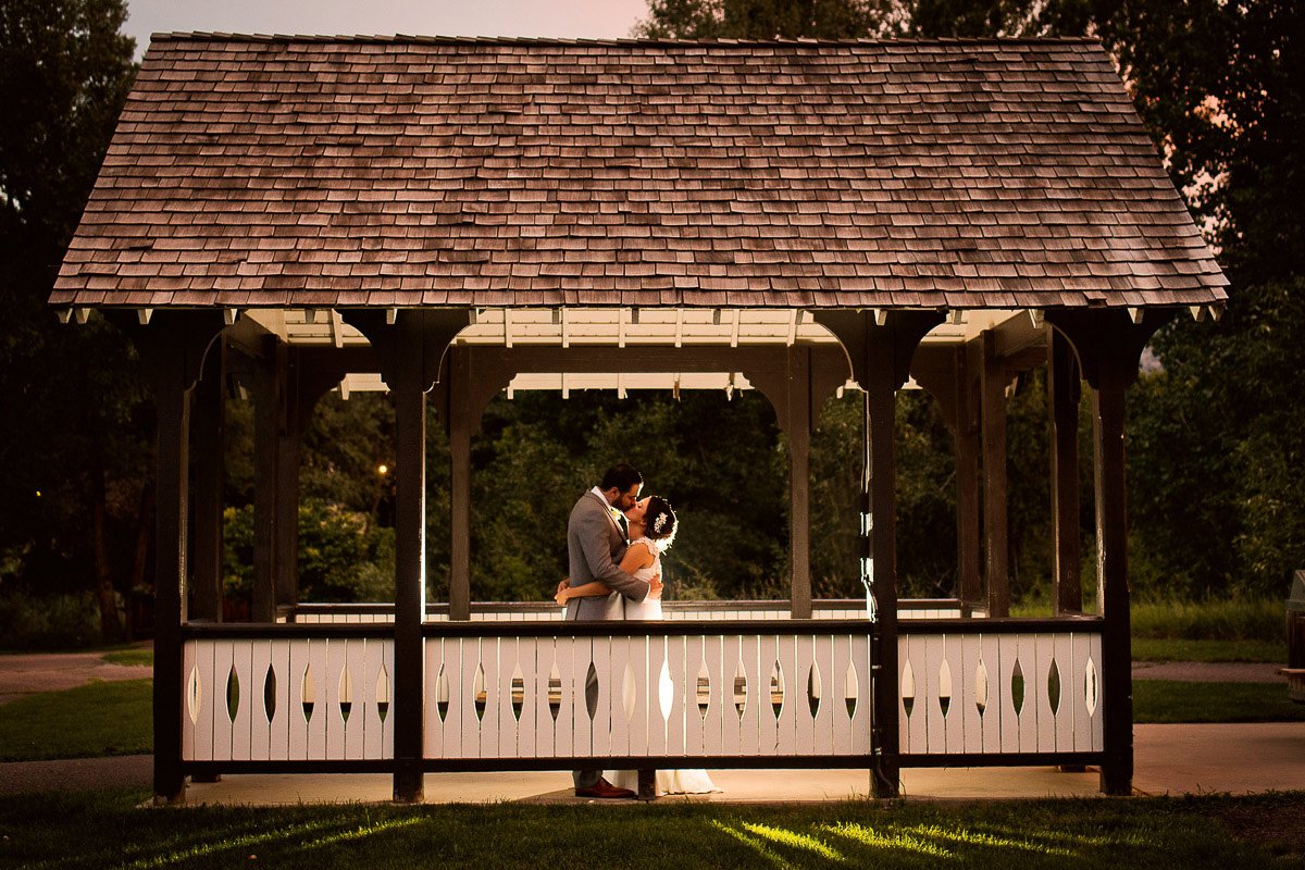 A couple embraces tenderly under a gazebo at dusk, during a Chatfield Farms wedding in Littleton, Colorado. The warm lighting casts a romantic glow, creating an intimate, serene mood.