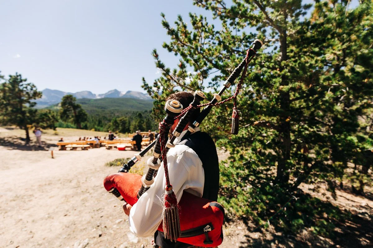 A bagpiper in traditional attire plays outdoors, with mountains in the background and seated guests in a sunny forest setting, creating a festive atmosphere.