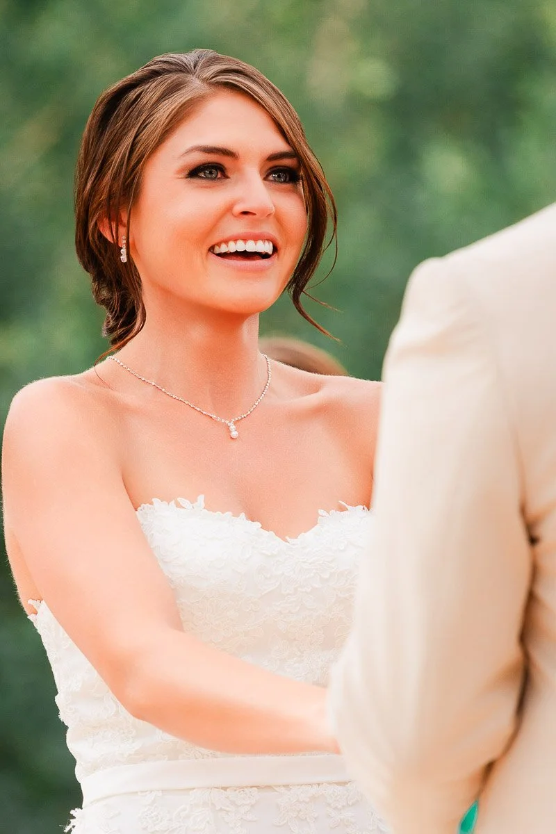 A bride in a lace wedding dress smiles brightly, exchanging vows outdoors. Her hair is elegantly styled, and she wears a delicate necklace. The mood is joyful.
