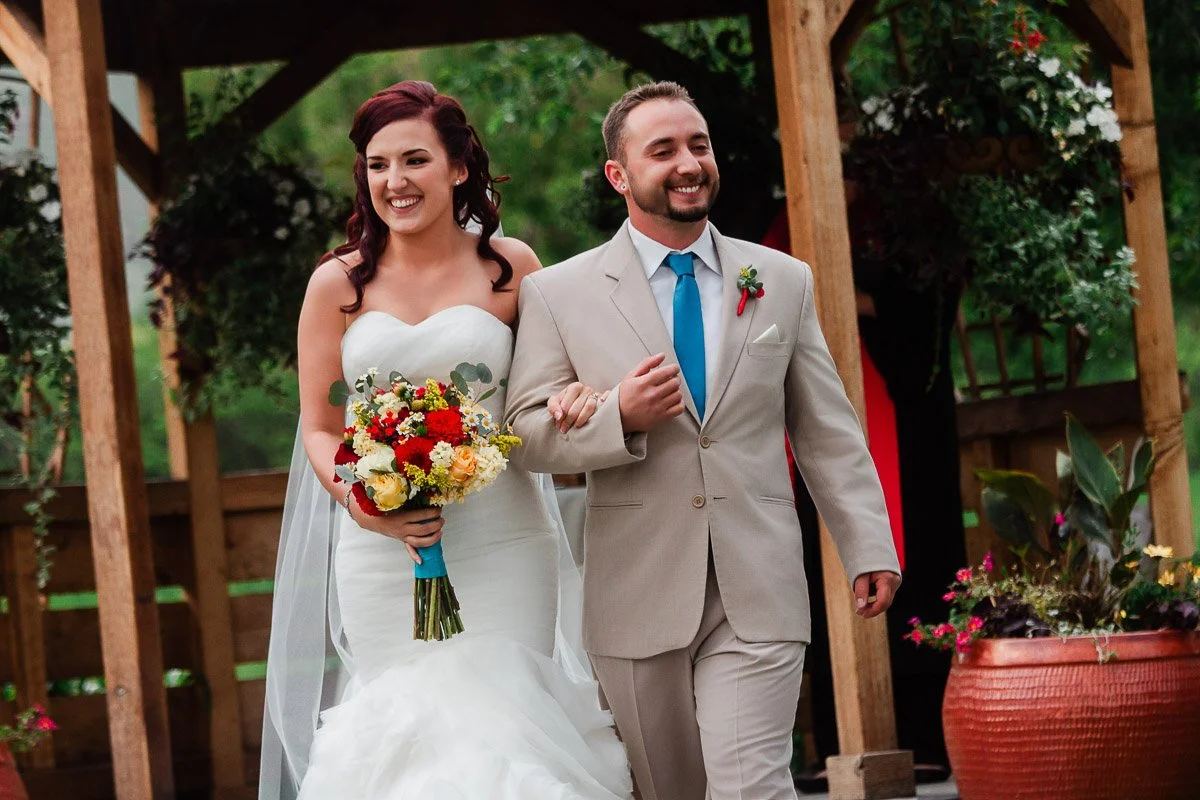 A bride in a white gown holds a vibrant bouquet, smiling alongside a groom in a beige suit with a blue tie. They walk joyfully under a wooden arch adorned with greenery.