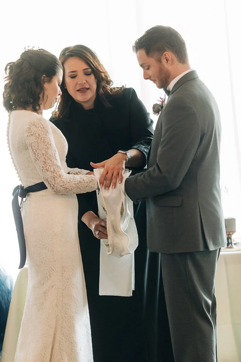 A couple stands facing each other during a wedding ceremony, holding hands wrapped with a cloth. A person in black officiates, creating a solemn and heartfelt atmosphere.