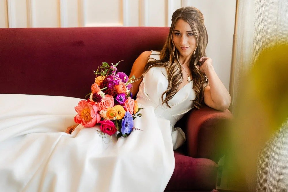 A woman in a white wedding dress reclines on a maroon sofa, holding a vibrant bouquet of pink, orange, and purple flowers, exuding elegance and joy.