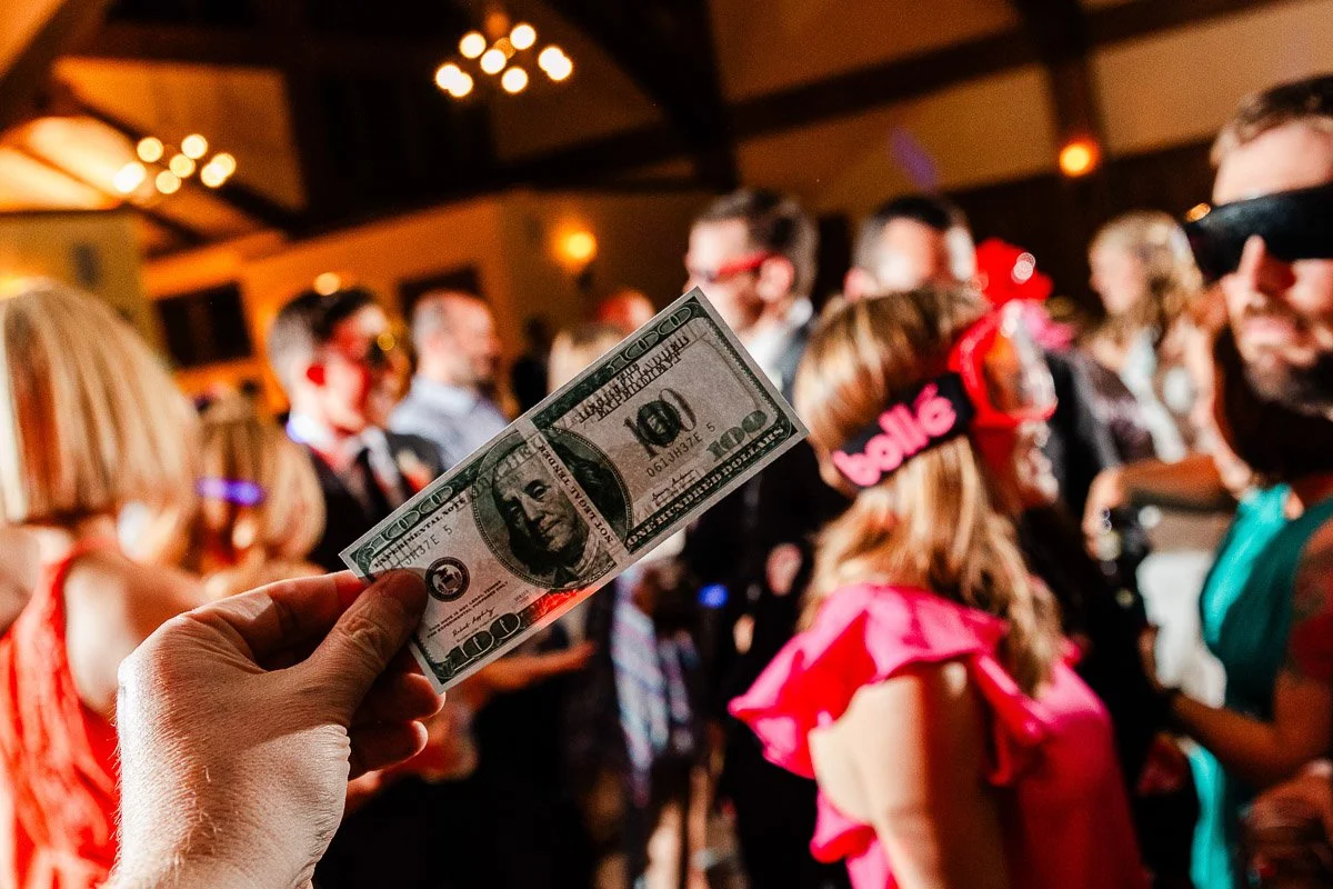 A hand holding a hundred-dollar bill in focus, with a lively party scene in the blurred background. Guests are wearing colorful, playful glasses.