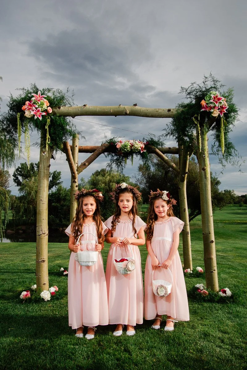 Three young flower girls in pink dresses and floral crowns stand under a wooden arch adorned with flowers, on a grassy field, creating a joyful, serene atmosphere during a Lakewood Country Club wedding.