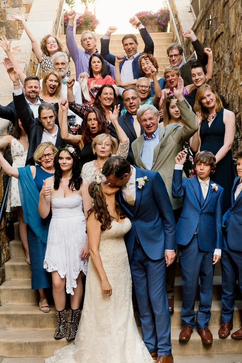 A joyful wedding group photo on stairs, with a bride and groom kissing in the center, surrounded by cheering family and friends with raised arms.