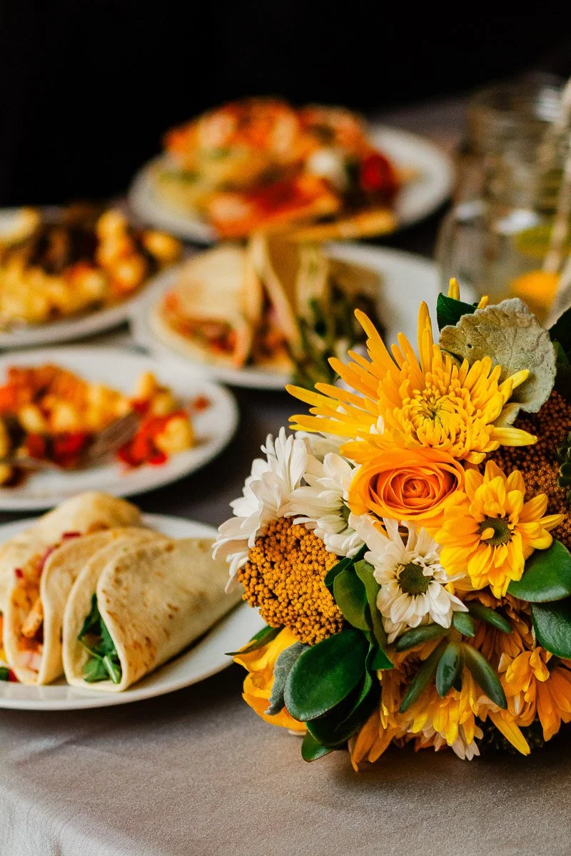 A vibrant bouquet of yellow and white flowers in the foreground, set against plates of assorted colorful food, creating a festive and warm atmosphere.