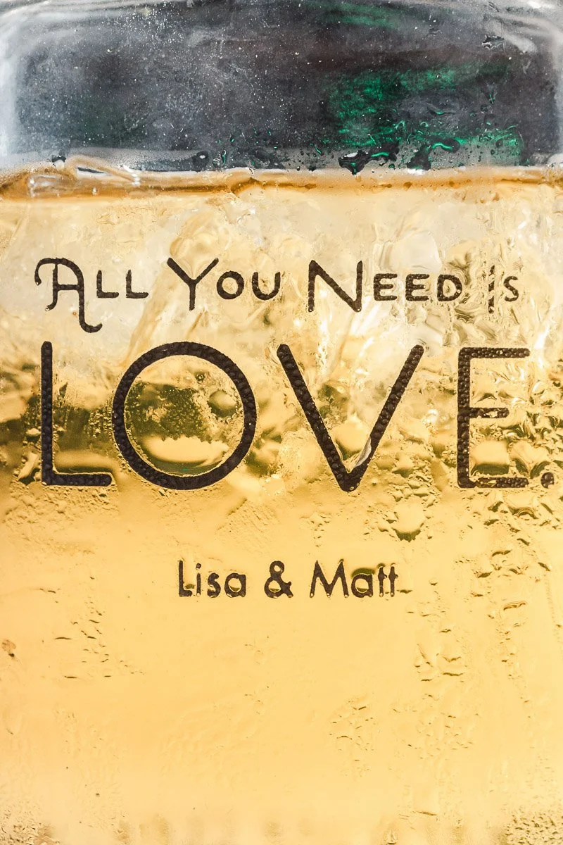 A close-up of a glass filled with a light amber liquid and ice. The phrase "All You Need is Love. Lisa & Matt" is printed in bold, dark lettering.