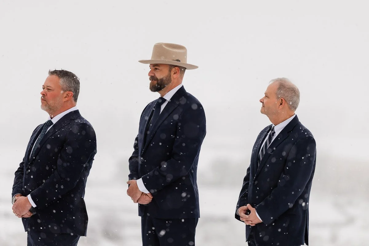 Three men in suits stand outdoors in the snow. The man in the center wears a beige hat. All appear focused and professional against a snowy backdrop.