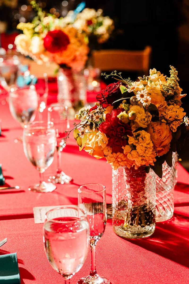 Elegant dining table setup with red cloth, featuring two bouquets of red and yellow flowers in glass vases, alongside shiny glassware, creating a warm ambiance.