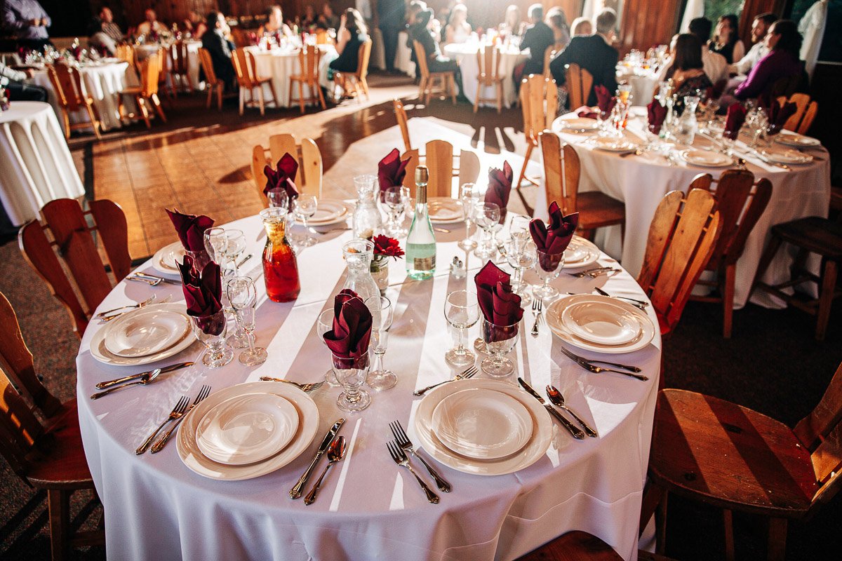 Elegantly set round tables with white linens, burgundy napkins, and sparkling glassware in a warmly lit banquet hall, creating a celebratory atmosphere.