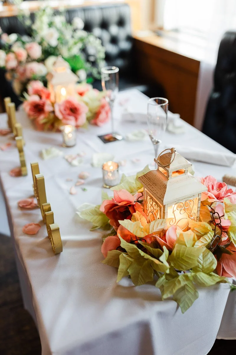 Elegant table setting with a white cloth, decorative lantern centerpiece surrounded by pink flowers and greenery. Soft candlelight creates a warm, romantic atmosphere.