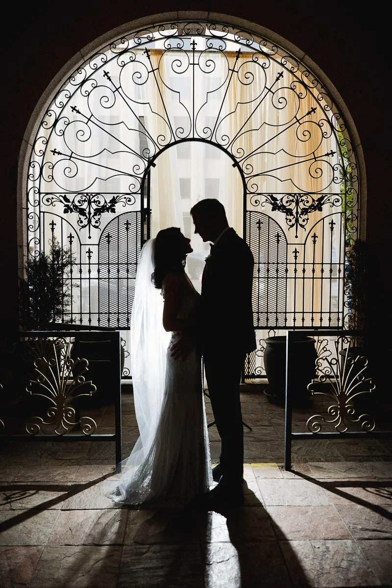 Silhouetted bride and groom embrace in front of ornate wrought-iron gate, backlit with soft light. Romantic and elegant wedding scene during a Mining Exchange Hotel wedding in Colorado Springs.