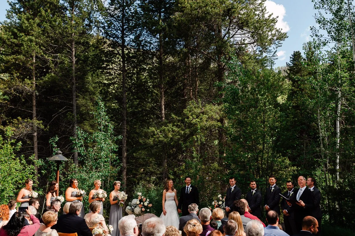Outdoor wedding ceremony with a bride and groom under tall trees. Bridesmaids and groomsmen stand beside them. Guests sit attentively, creating a serene atmosphere.