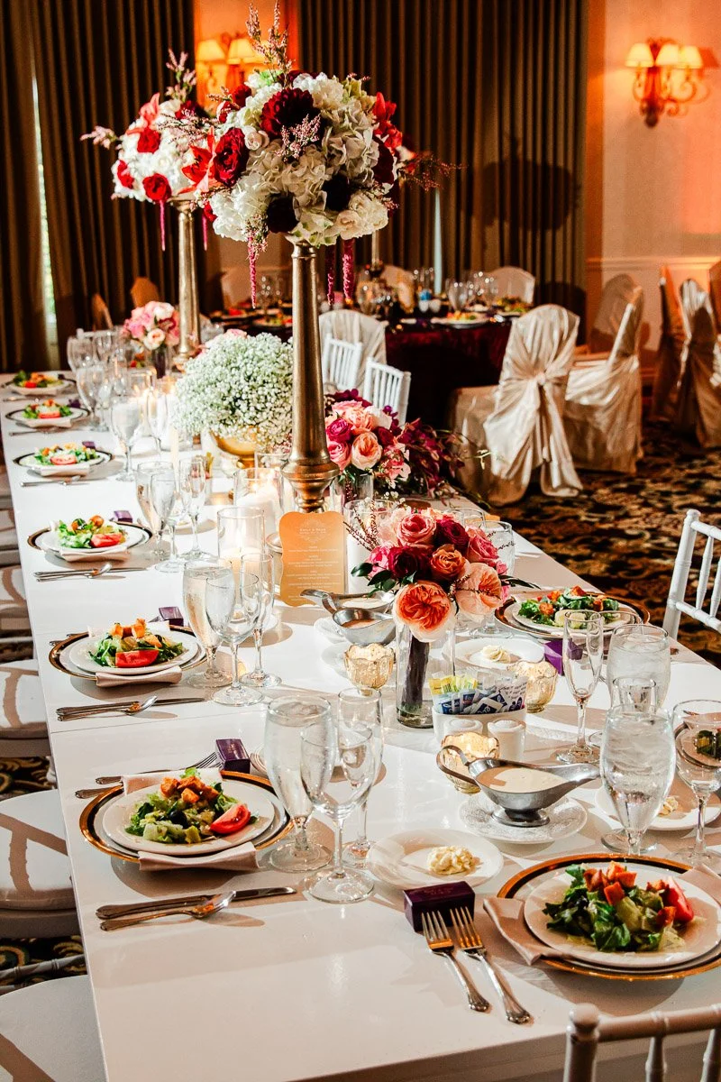Elegant banquet setup with a long white table adorned with floral centerpieces and candles. Plates of vibrant salads and crystal glassware enhance the luxurious ambiance.
