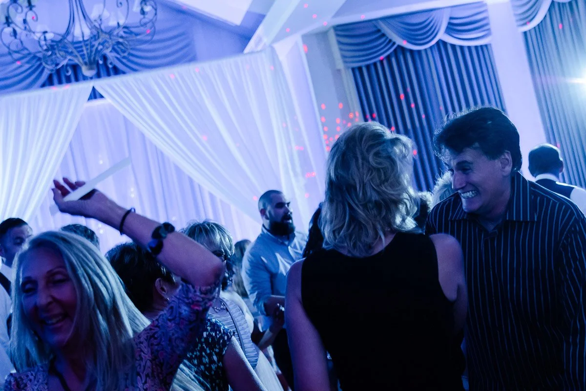 A lively party scene with guests dancing under blue lighting. A woman in a patterned dress smiles joyfully. Draped curtains and chandeliers enhance the festive mood.