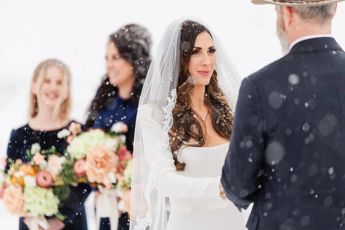 Bride in a white gown and veil smiles tenderly at groom during snowy outdoor wedding ceremony. Bridesmaids in dark dresses hold colorful bouquets.
