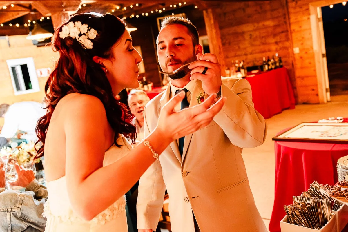 A couple in wedding attire is playfully eating cake inside a warmly lit rustic venue. Their expressions are joyful and lighthearted, enhancing the festive atmosphere.