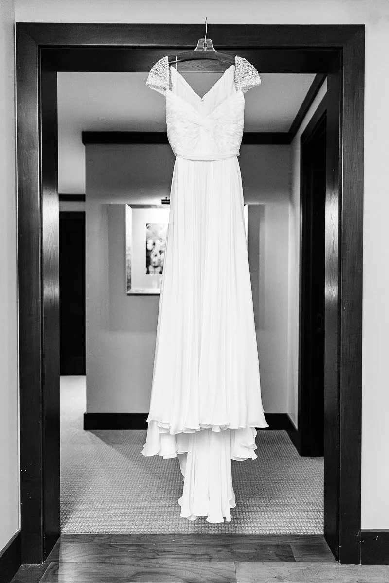 A white wedding dress with lace cap sleeves hangs elegantly in a doorway. The setting is softly lit, conveying a serene and anticipatory mood.