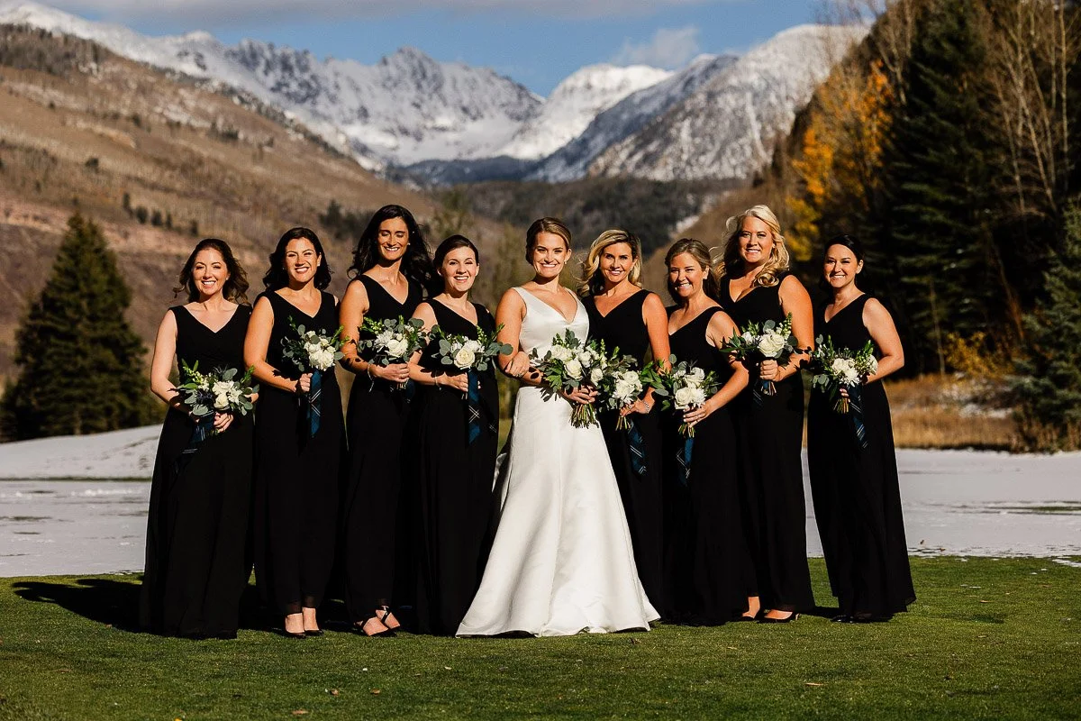 A bride in a white gown stands with eight bridesmaids in black dresses holding bouquets. They are outdoors with a snowy mountain backdrop and greenery.